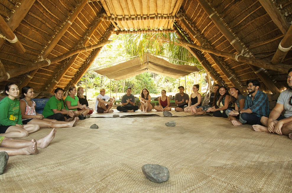 Retreat-Gruppe sitzt im Kreis in traditionellem Hawaiianischem Versammlungshaus, umgeben von tropischer Vegetation
